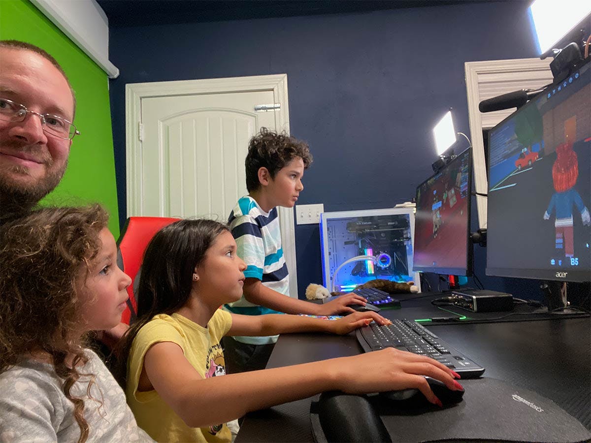 Photo of Mission Mike playing videogames with his three children. Sitting at a computer desk with a green screen behind them.